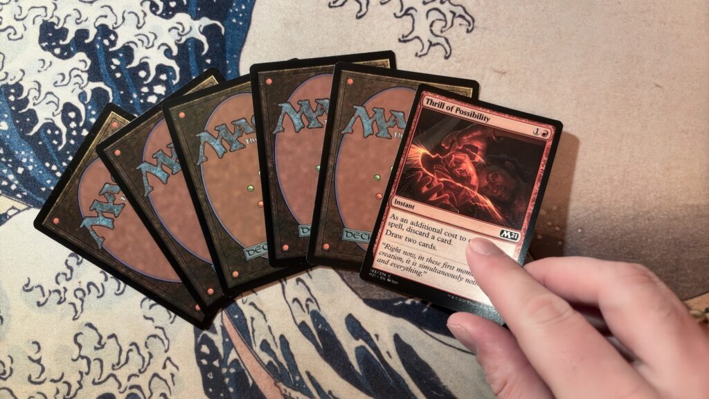 Eine Person hält die "Thrill of Possibility"-Karte aus dem Sammelkartenspiel Magic: The Gathering in der Hand. Die Karte ist rot umrandet und hat einen Instant-Effekt, der das Ziehen von zwei Karten ermöglicht, nachdem eine Karte abgeworfen wurde. Im Hintergrund liegen fünf weitere Magic-Karten mit der Rückseite nach oben aufgefächert auf einer Spielmatte mit einem Design, das an die berühmte japanische Welle von Hokusai erinnert.