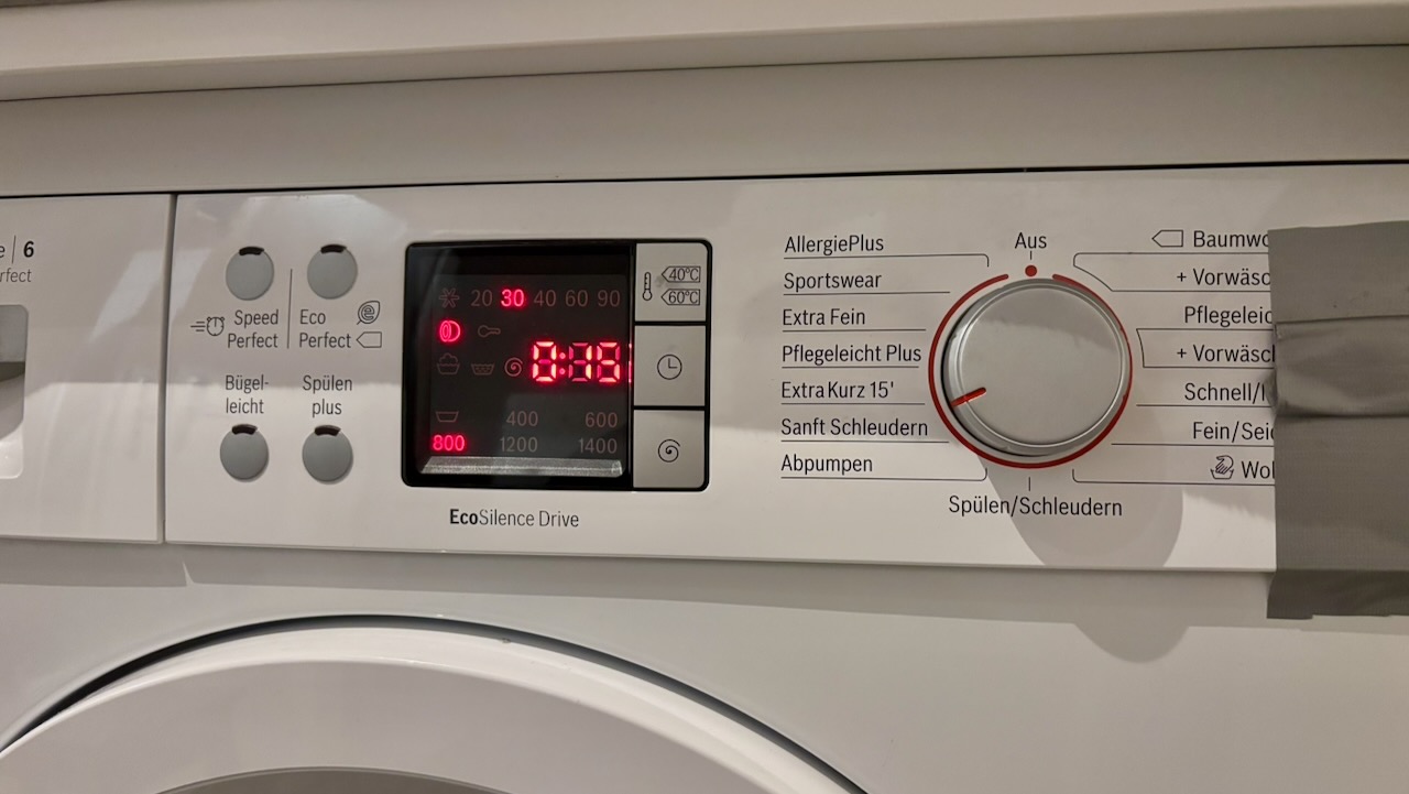 Frontansicht einer Waschmaschine. Das Bedienfeld zeigt ein digitales Display mit rot leuchtender Restzeit „0:38“ und mehreren Symbolen für Waschprogramme. Rechts befindet sich ein Drehregler für verschiedene Programme wie „AllergiePlus“, „Sportswear“, „Extra Fein“ und „Sanft Schleudern“. Einige Funktionstasten links des Displays sind beschriftet mit „Bügel-leicht“, „Spülen plus“, „Speed Perfect“ und „Eco Perfect“. Auf der rechten Seite ist ein Stück Klebeband zu sehen.