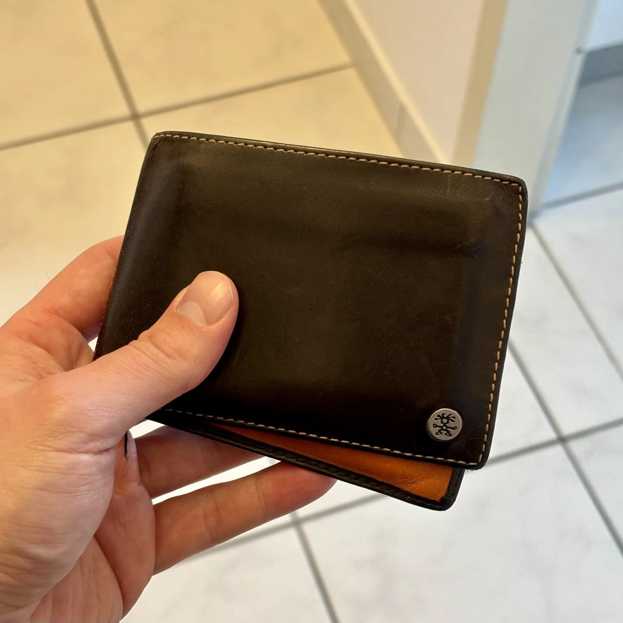ine Hand hält eine schwarze Leder-Geldbörse mit orangefarbenem Innenfutter. Auf der Vorderseite ist ein metallisches Crumpler Logo eingearbeitet.