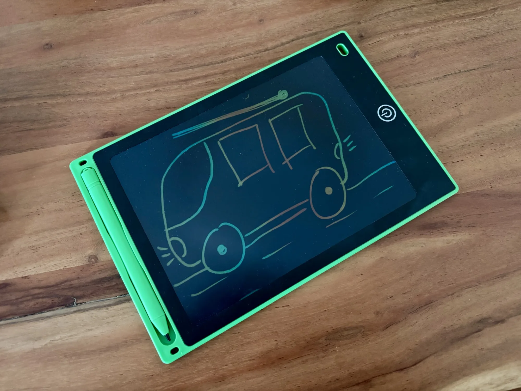 Ein grünes LCD-Zeichenbrett liegt auf einem Holztisch. Auf dem Display ist eine einfache Zeichnung einer Feuerwehr mit Rädern und Fenstern zu sehen. Das Zeichenbrett hat seitlich einen Stift in einer Halterung.