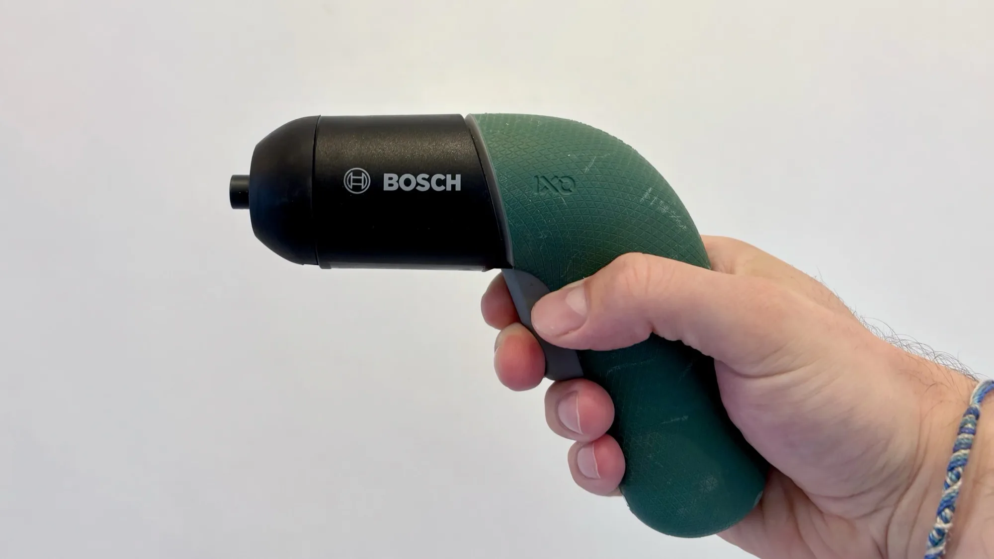 Eine Person hält einen grünen Bosch IXO 6 Akkuschrauber seitlich in der rechten Hand vor einer schlichten weißen Wand. Das kompakte Werkzeug hat ein minimalistisches, abgerundetes Design mit schwarzem Vorderteil und grünem Griff. Das Bosch-Logo ist gut sichtbar auf der Gehäuseseite. Der Schrauber ist ausgeschaltet und ohne eingesetzten Bit fotografiert.
