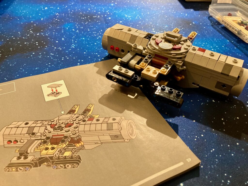 Ein angefangenes LEGO Millennium Falcon Modell ist auf einer künstlerischen Darstellung eines Sternenhimmels positioniert, um zu simulieren, dass es durch den Weltraum fliegt. Die Perspektive ist so gewählt, dass das Modell dynamisch und dreidimensional erscheint, mit dem Fokus auf der Vorderseite und den Seiten des Schiffes.