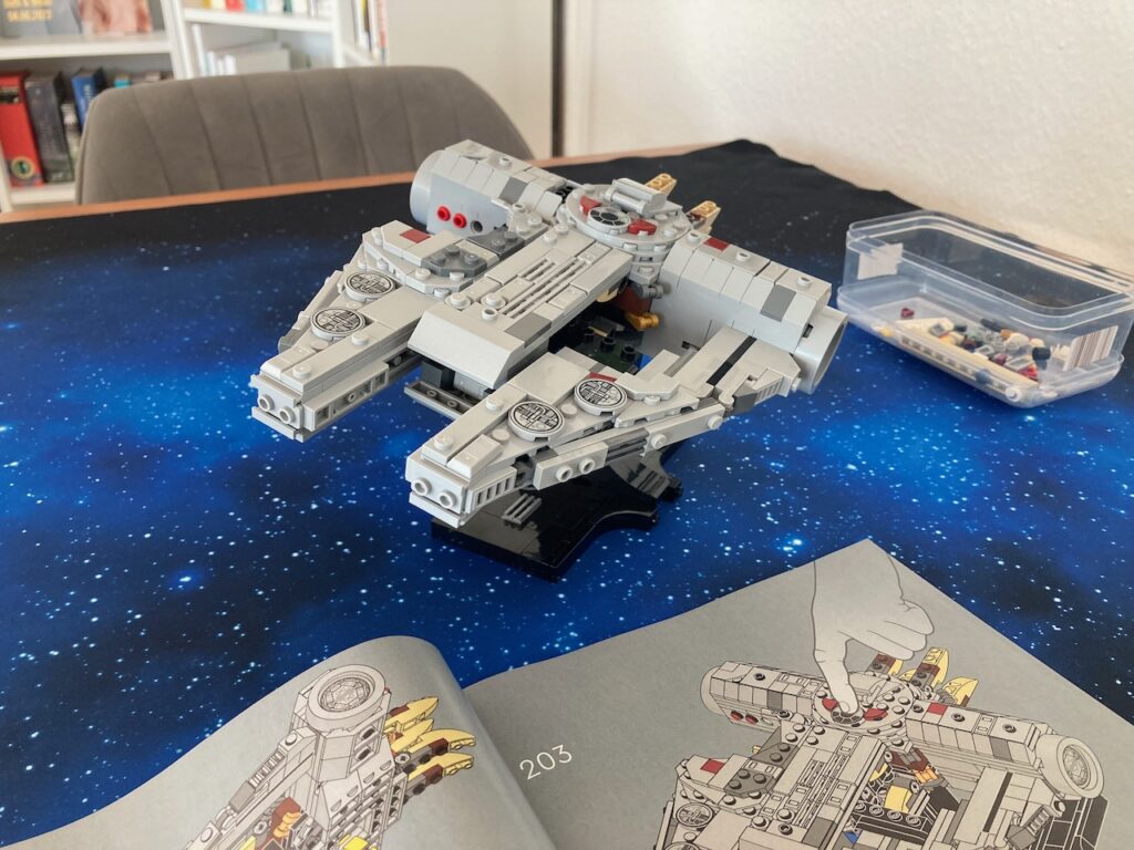 Der halb aufgebaute LEGO Millennium Falcon auf einer grauen Unterlage, leicht von oben fotografiert. Das Modell ist detailliert und zeigt verschiedene Farben, hauptsächlich in Weiß und Grau.