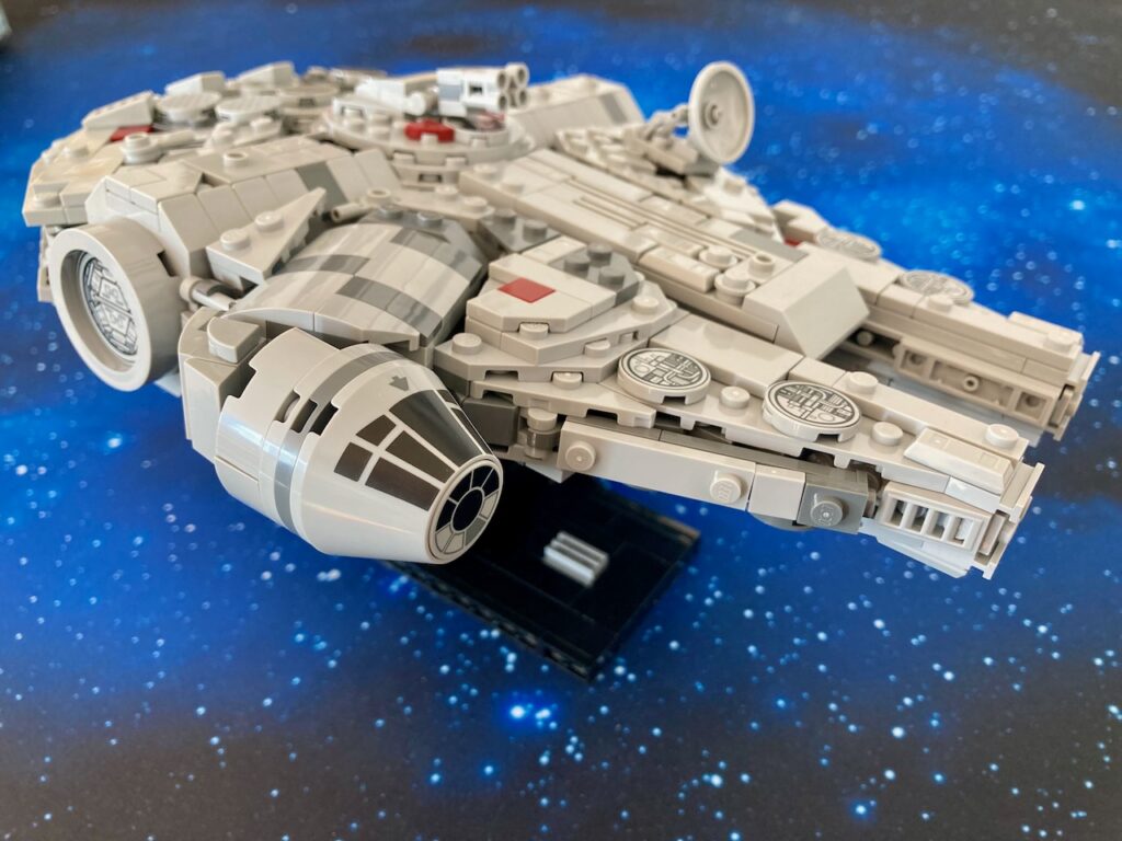 Eine Nahaufnahme des LEGO Millennium Falcon, der auf einer Unterlage mit Sternenhimmel-Muster steht, was den Eindruck erweckt, dass das Raumschiff im Weltraum schwebt. Das Modell ist im Detail sichtbar, einschließlich der runden Schüsseln und der roten Akzente.