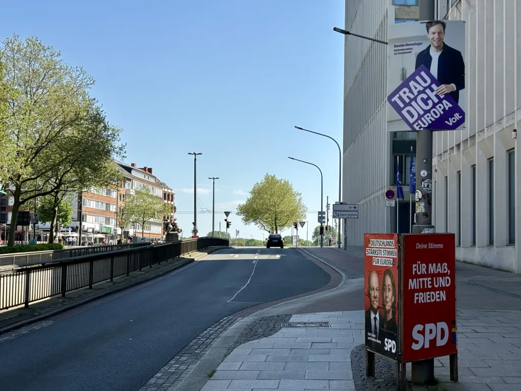 Städtische Straßenszene mit politischen Wahlkampfplakaten an einem Laternenpfahl und Schildern am Straßenrand, klarem blauem Himmel und wenig Verkehr.