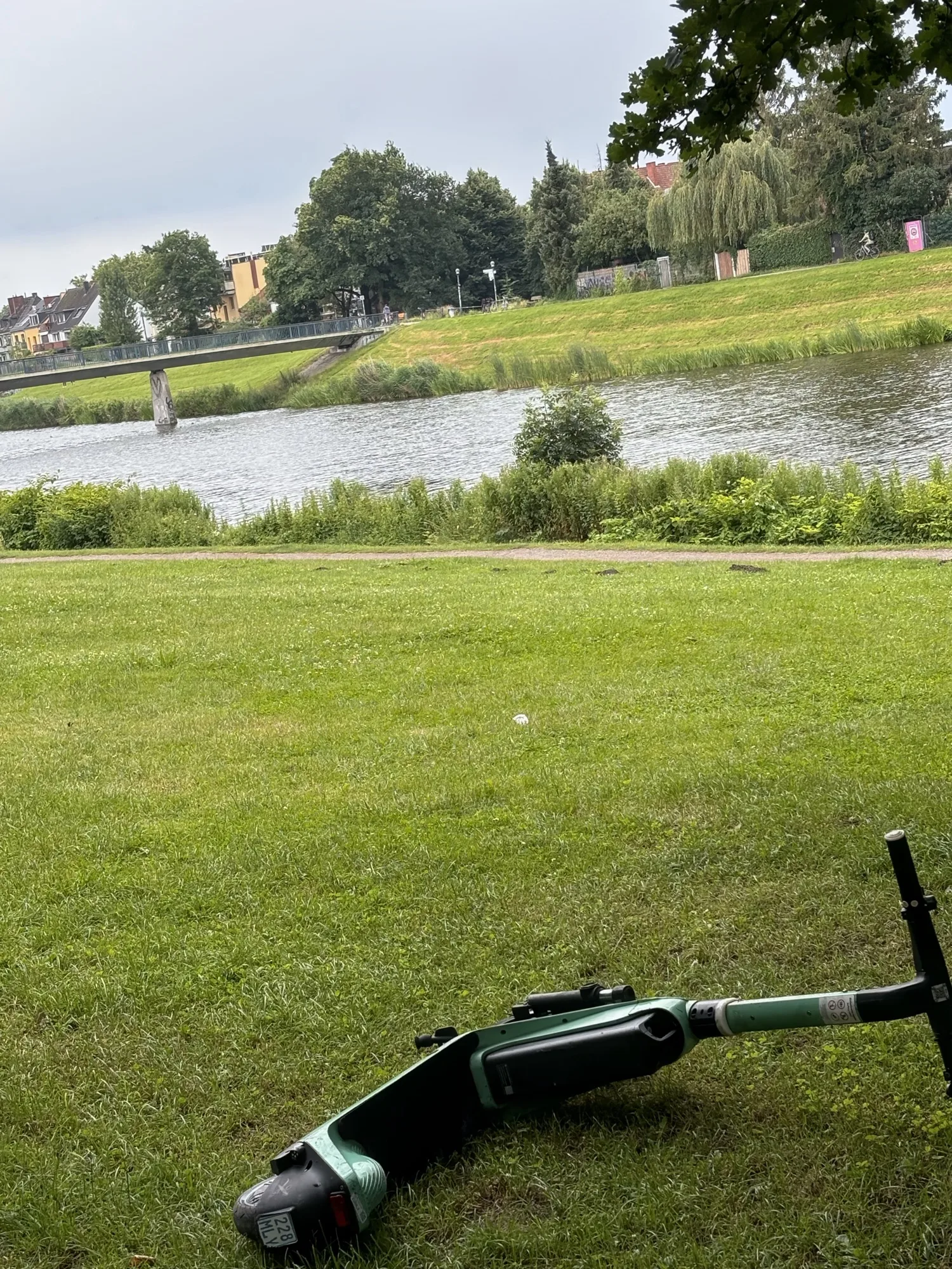 Dieses Bild zeigt eine Parklandschaft mit einem Fluss im Hintergrund, über den eine Fußgängerbrücke führt. Im Vordergrund liegt ein grüner Elektroroller auf dem Gras. Am anderen Ufer des Flusses sind Bäume und Häuser zu sehen. Das Bild vermittelt eine ruhige und friedliche Atmosphäre.