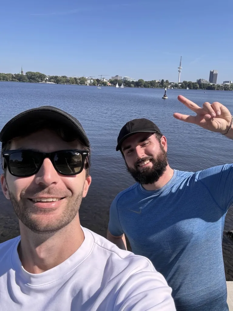 Zwei bärtige Männer mit Mütze schauen in die Kamera und lächeln. Der vordere Mann hat ein weißes Shirt und eine Sonnenbrille. Der hintere Mann ein blaues Sportshirt und macht das Victory Zeichen. Sie stehen vor Wasser. Im Hintergrund sieht man Teile von Hamburg.