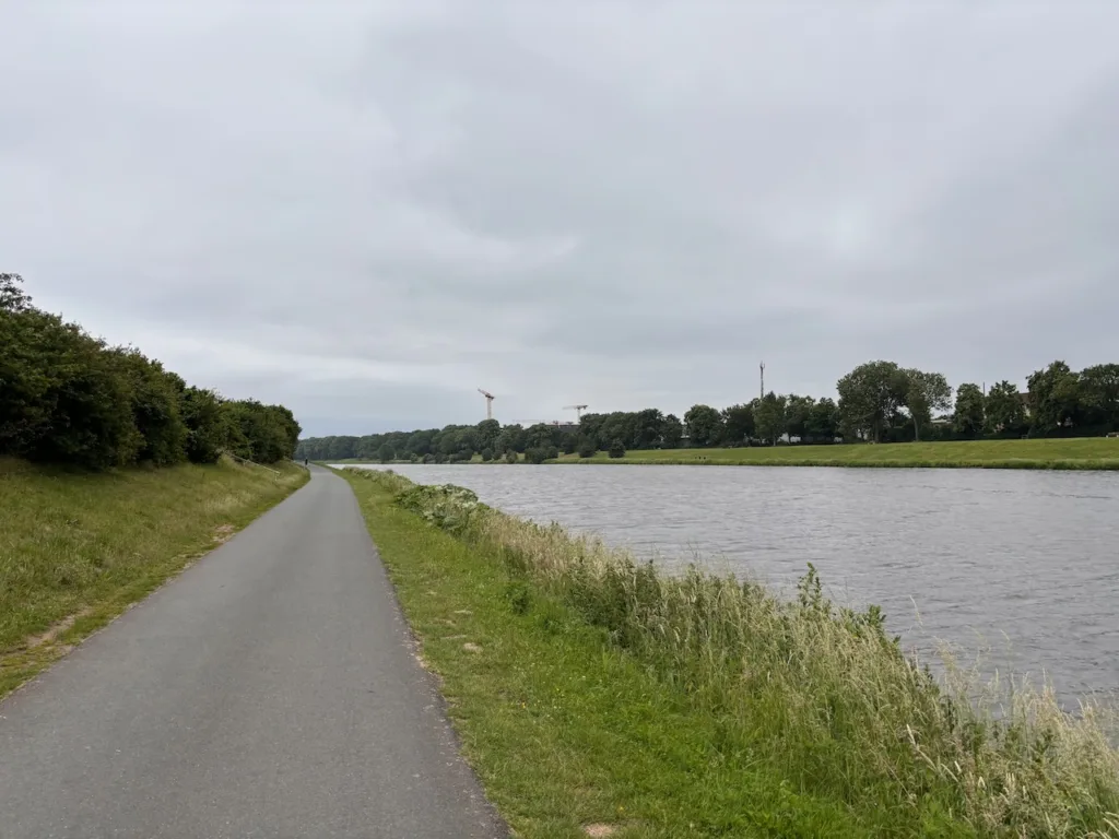 Ein asphaltierter Weg an einem Fluss. Im Hintergrund sieht man Bäume und Baukräne.