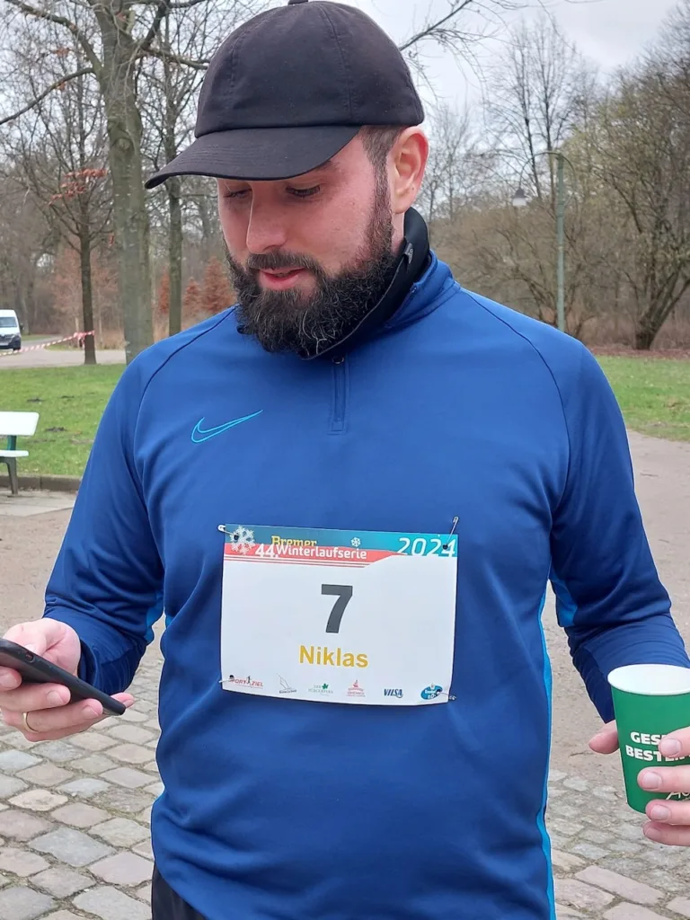 Ein bärtiger Mann in einem dunkelblauen Langarm-Laufshirt und schwarzer Mütze schaut auf sein Handy. Auf seinem Shirt trägt er eine Startnummer mit der Nummer 7 und dem Namen „Niklas“. In seiner Hand hält er einen grünen Becher. Im Hintergrund sieht man eine parkähnliche Umgebung mit Bäumen und Kopfsteinpflasterwegen. Das Wetter wirkt kühl und bedeckt.