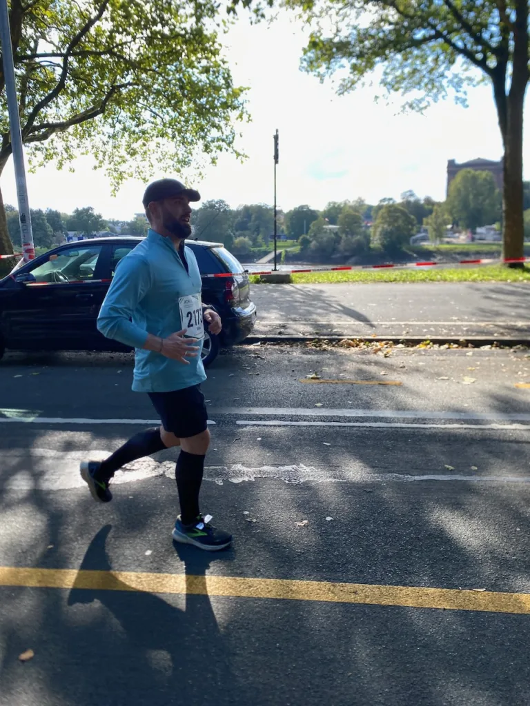 Ein Mann läuft draußen entlang einer von Bäumen gesäumten Straße, gekleidet in ein hellblaues Langarmshirt, schwarze Shorts, kniehohe Socken und Laufschuhe. Er wirkt konzentriert und hat ein leichtes Lächeln, während er an einem Rennen teilnimmt. Im Hintergrund sind Autos und Bäume unter klarem Himmel zu sehen.